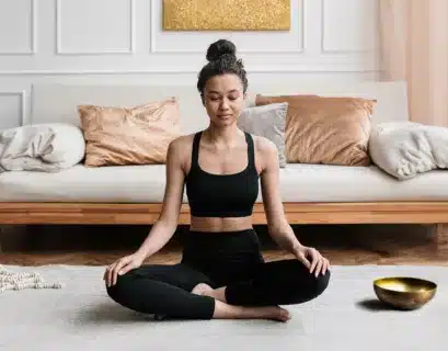 Frau sitzt im Meditationssitz auf dem Teppich vor einer Couch und macht eine Vipassana-Meditation