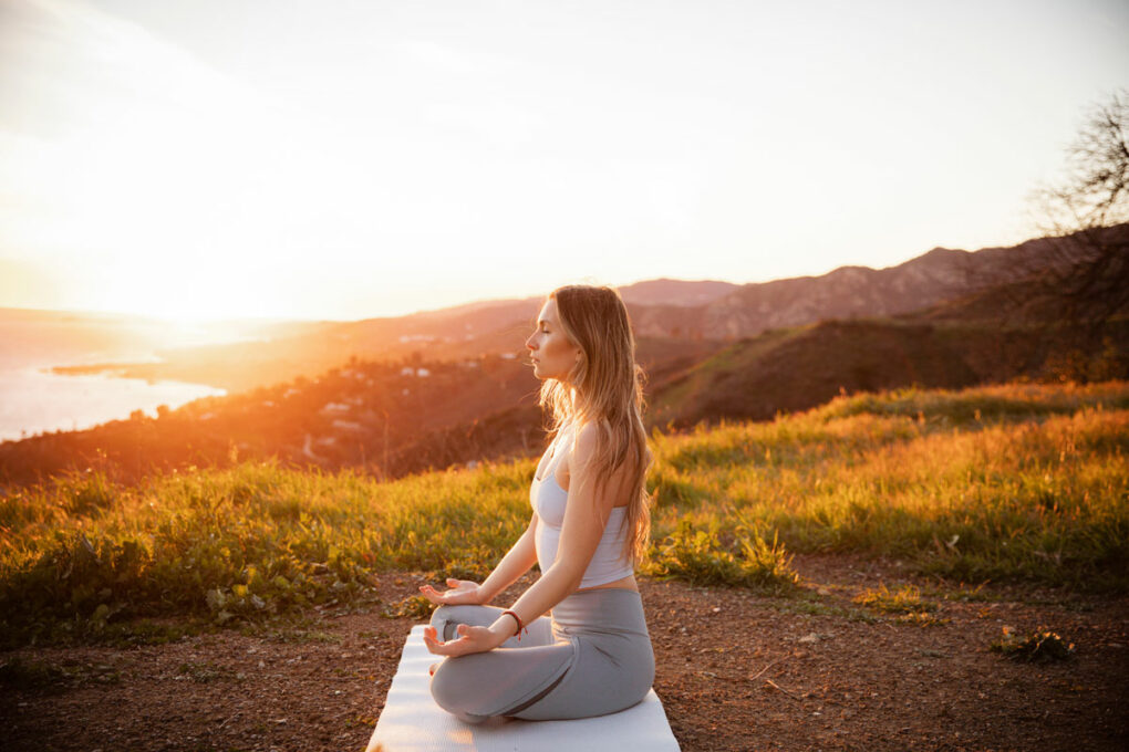 Seitenansicht einer meditierenden Sportlerin, die auf einer Übungsmatte sitzt, während die Sonne untergeht