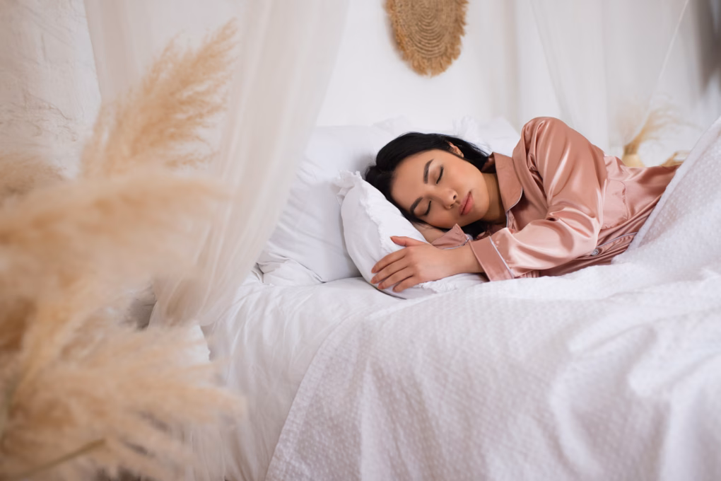 Hübsche junge asiatische Frau in Seidenpyjama schläft im Bett mit weißer Bettwäsche im Schlafzimmer.