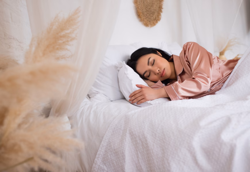 Hübsche junge asiatische Frau in Seidenpyjama schläft im Bett mit weißer Bettwäsche im Schlafzimmer.