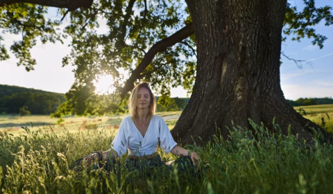 Frau sitzt in der Natur vor einem Baum und meditiert
