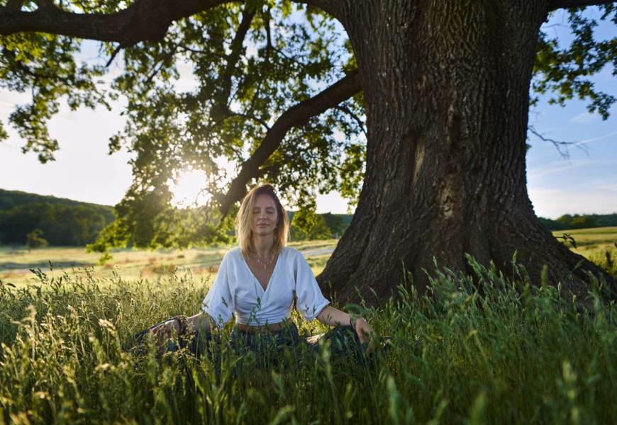 Frau sitzt in der Natur vor einem Baum und meditiert