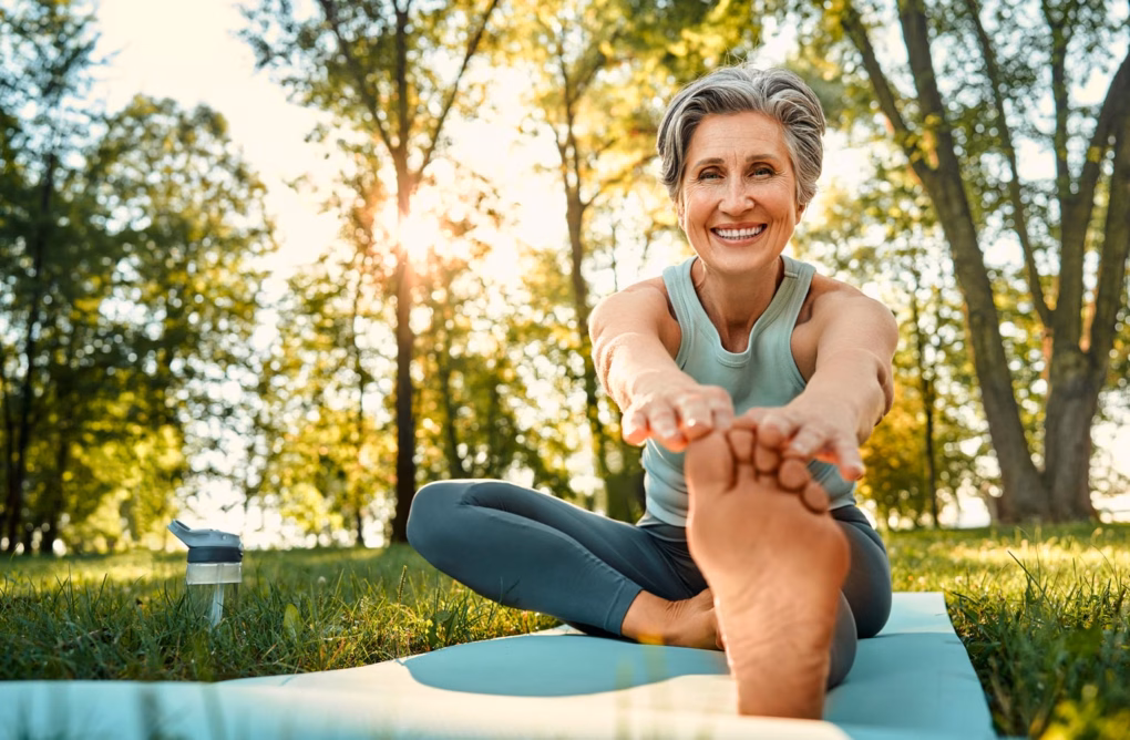 Porträt einer fröhlichen Frau mit trendiger Frisur, die im Freien auf einer Yogamatte trainiert und nach ihren Zehen greift.