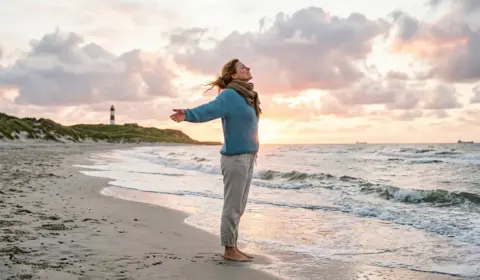 Entpannte Frau am Strand bei Sonnenaufgang, die tief einatmet und die Heilkraft der Klimatherapie genießt.
