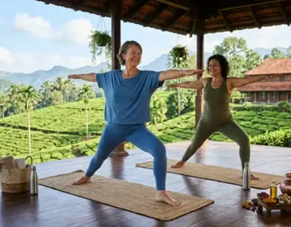 Zwei Frauen über 50 praktizieren Yoga in der Krieger-Pose während eines Ayurveda-Retreats in einer tropischen Hügellandschaft.