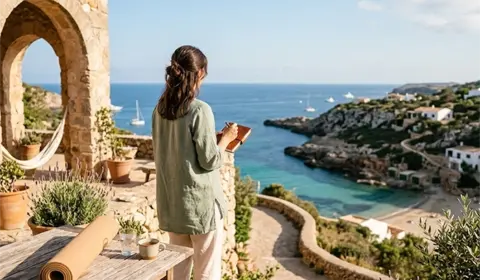 Eine Frau steht barfuß auf einer sonnigen Terrasse mit Blick auf das blaue Meer, schreibt in ein Notizbuch; im Vordergrund liegt eine zusammengerollte Yogamatte aus Kork auf einem Holztisch.